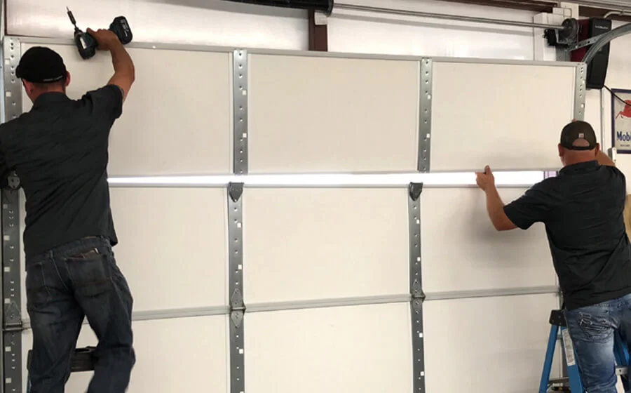 Two workers installing a white sectional garage door, with one using a power drill to secure a panel.