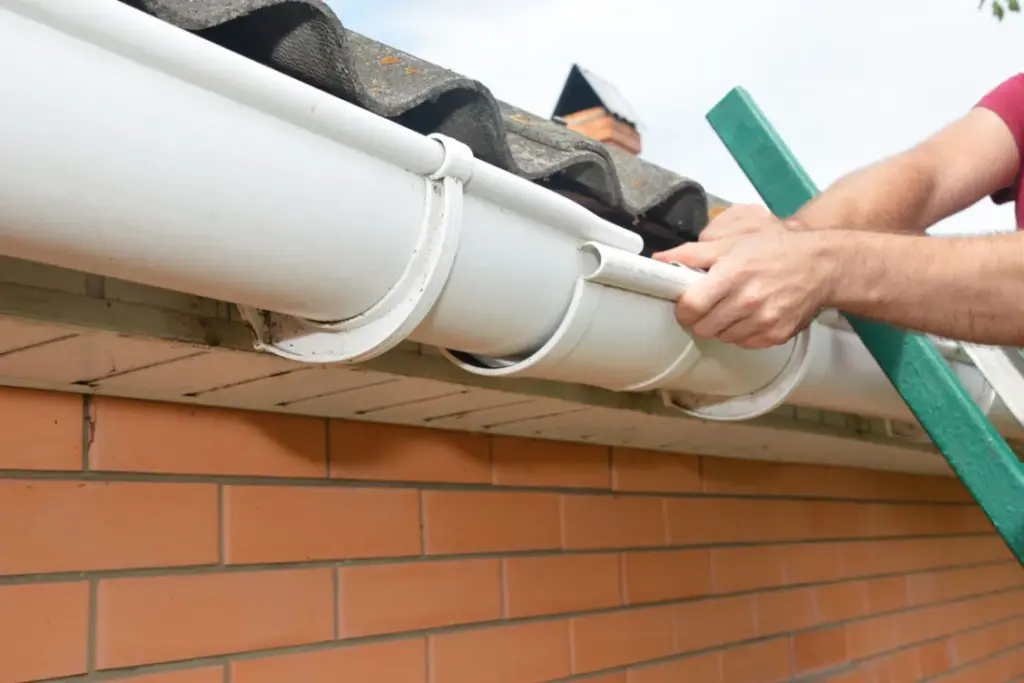Hands installing white plastic gutters on the eaves of a brick house.
