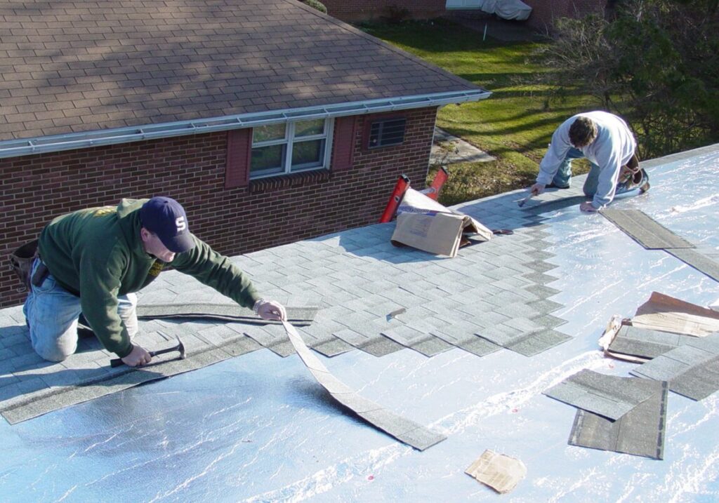 Two workers installing grey asphalt shingles on a house roof over silver underlayment.
