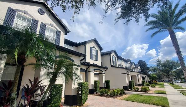 Two-story white townhomes with dark shutters, palm trees, and a sidewalk under a blue sky with clouds.