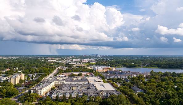 Aerial view of suburban buildings, greenery, and a lake, with a distant city skyline under dramatic storm clouds.
