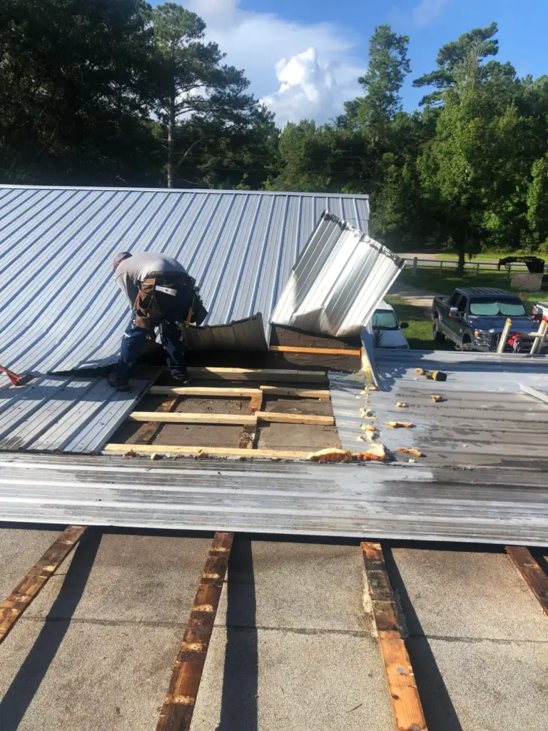 Worker in a tool belt removing corrugated metal roofing panels, exposing the wooden support beams.