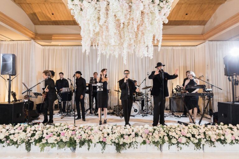 A live band in black attire performs on a stage decorated with flowers under a large white floral chandelier.
