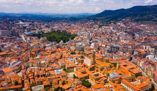 Panoramic aerial view of a city with orange rooftops, a cathedral, a green park, and mountains in the background.