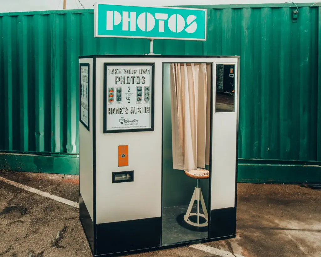 A white and black Photo-matica photo booth with a tan curtain and stool, set against a green shipping container.
