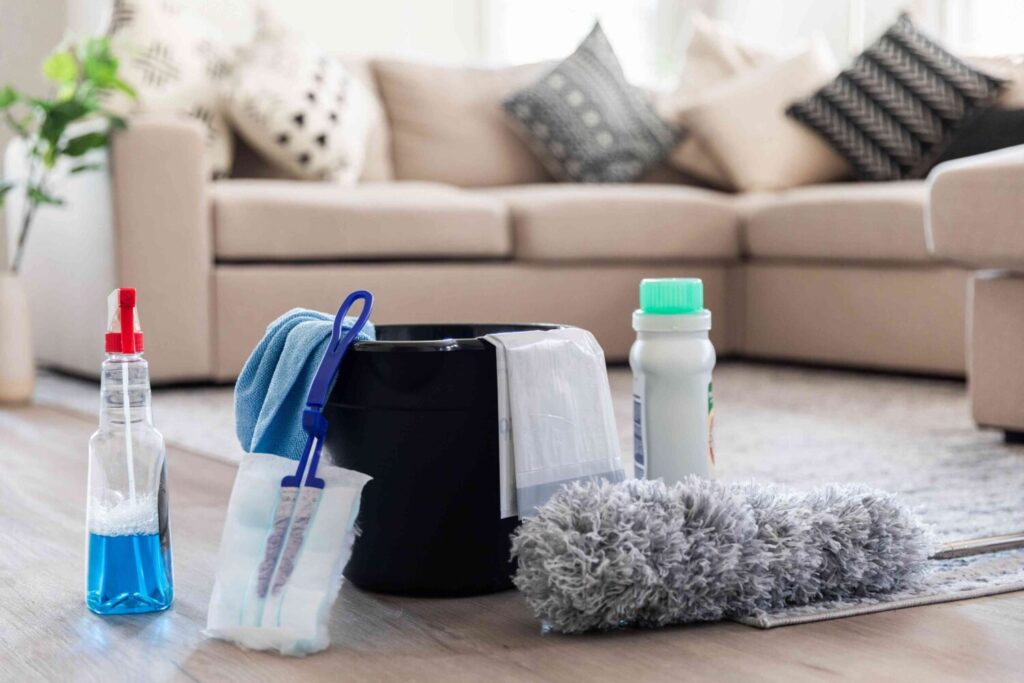 Cleaning supplies including a bucket, spray bottle, and microfiber duster on a living room floor.