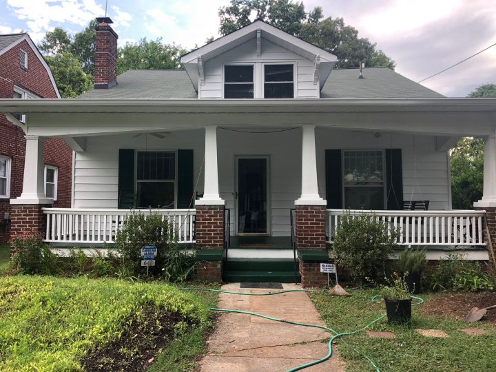 A white single-story bungalow with a brick-pillared front porch, a gabled roof dormer, and a brick chimney.