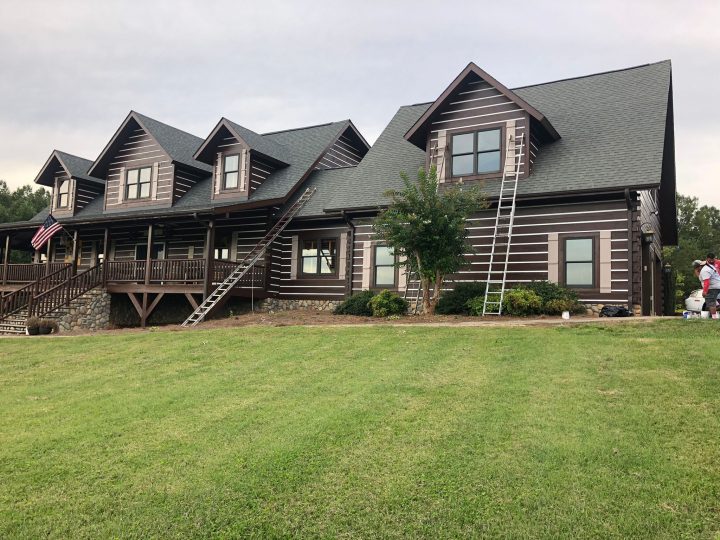 Large multi-story log cabin home with ladders against the exterior for maintenance or painting, set on a grassy lawn.