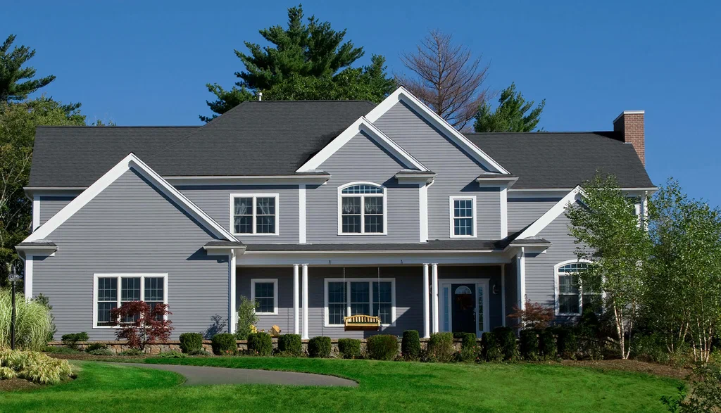 Large gray two-story suburban house with a porch swing, white trim, and a manicured lawn under a clear blue sky.