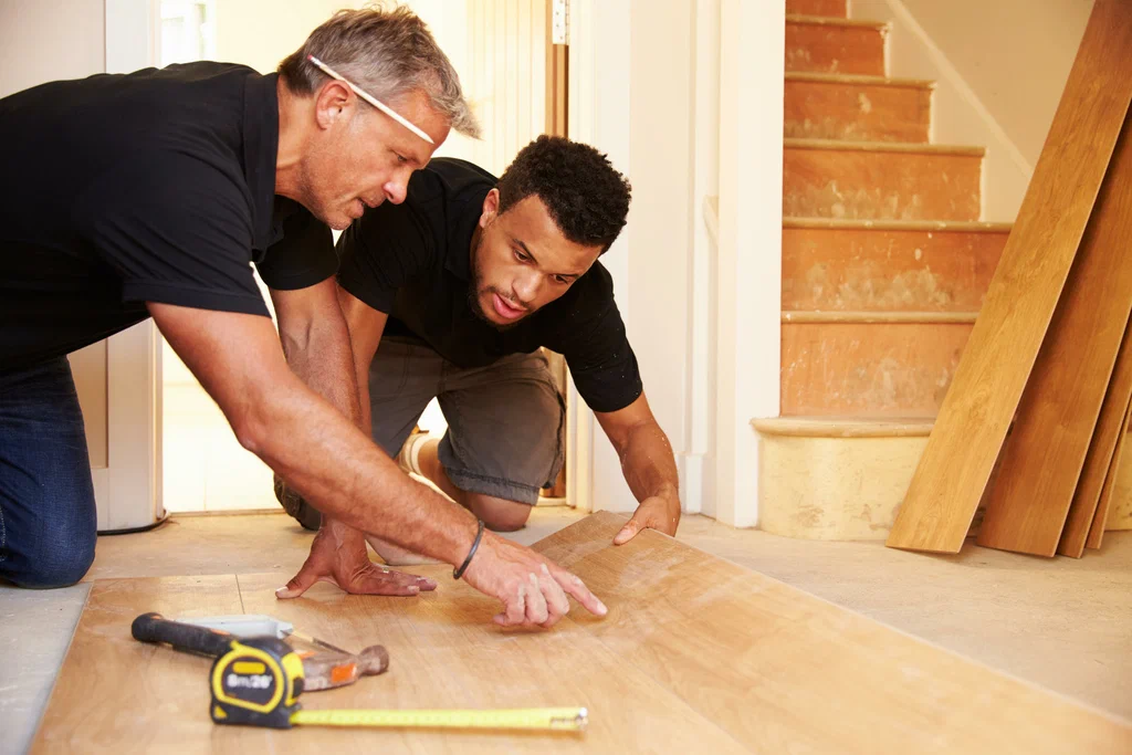 Two men installing wood flooring, examining a plank with tools nearby.