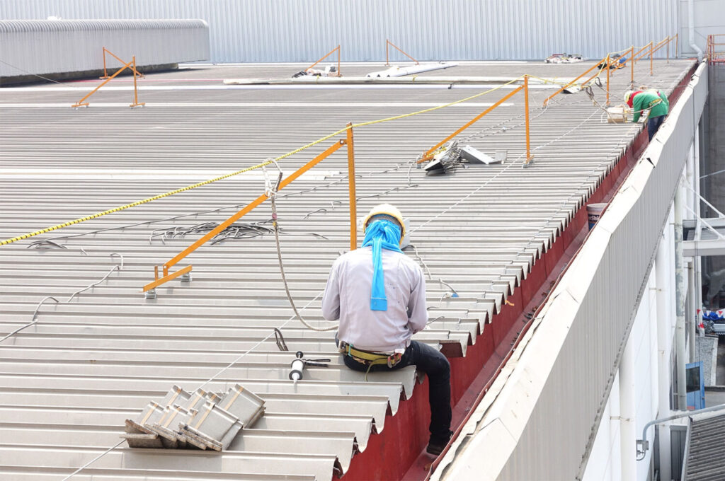 Workers on a corrugated metal roof wearing safety harnesses attached to yellow fall protection railings.