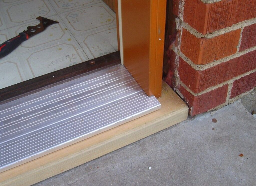 A metal threshold installed on a wooden sill at an exterior doorway with a brick wall and a floor scraper nearby.