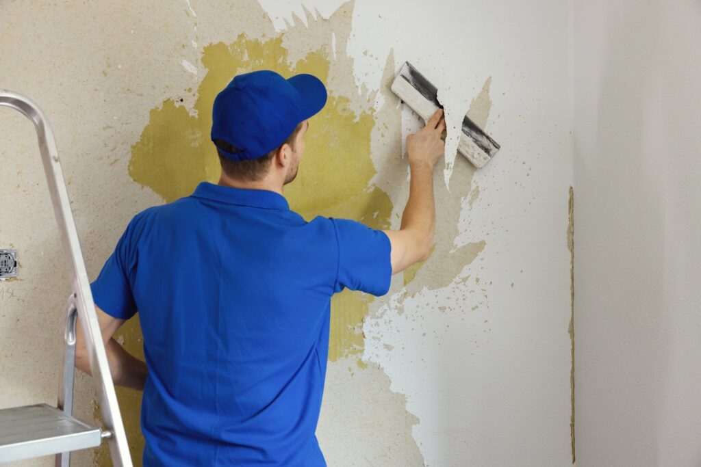 Worker in blue uniform scraping old wallpaper off a wall with a putty knife.