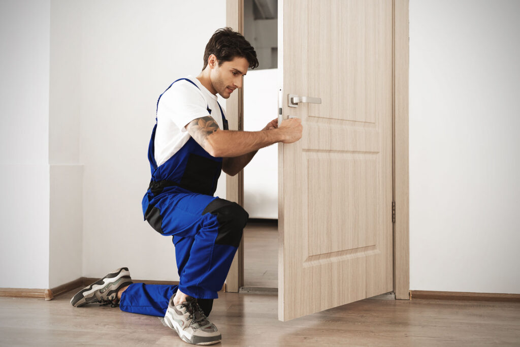 Handyman in blue workwear kneeling to install a metal handle on a wooden door.