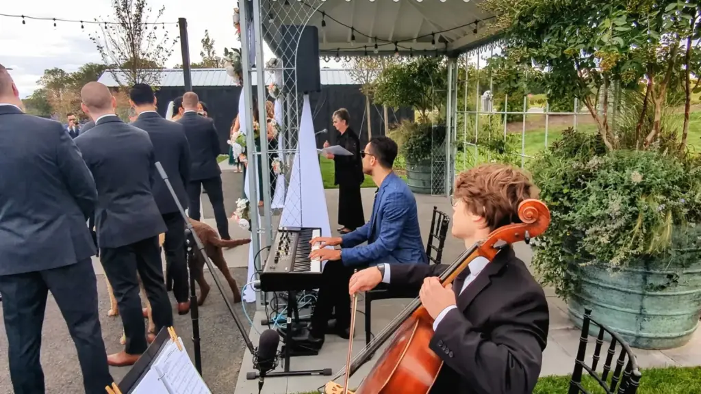 Musicians play cello and keyboard during an outdoor wedding ceremony under a pavilion.