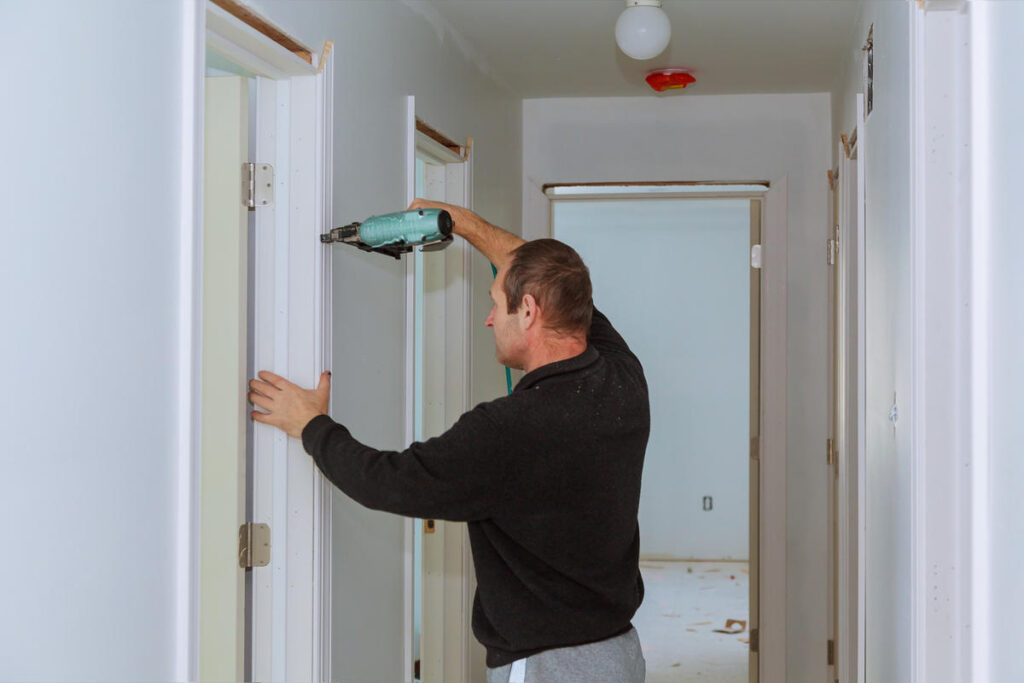 A carpenter uses a nail gun to install wooden door trim in a hallway.