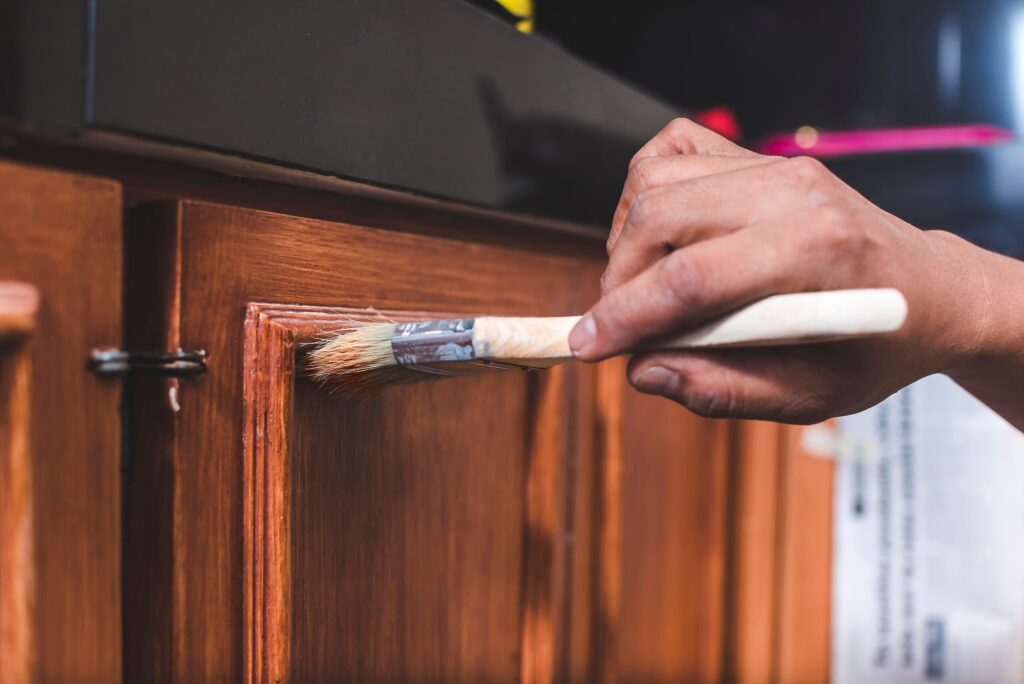 Close-up of a person's hand using a brush to apply wood stain to a cabinet door.