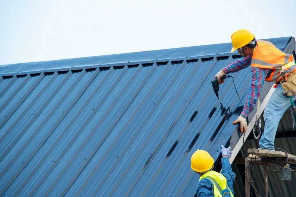 Construction workers in safety gear installing a blue corrugated metal roof using a power drill.