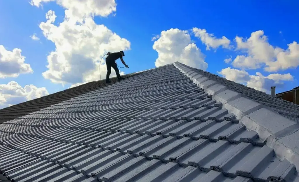 Worker spraying a grey tiled roof under a bright blue sky with clouds.