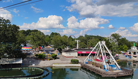Amusement park with a boat swing ride on a pond, colorful carousel-style rides, and trees under a blue sky with clouds.