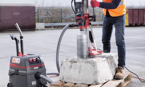Worker using a diamond core drill on a concrete block, connected to an AGP vacuum extraction system.