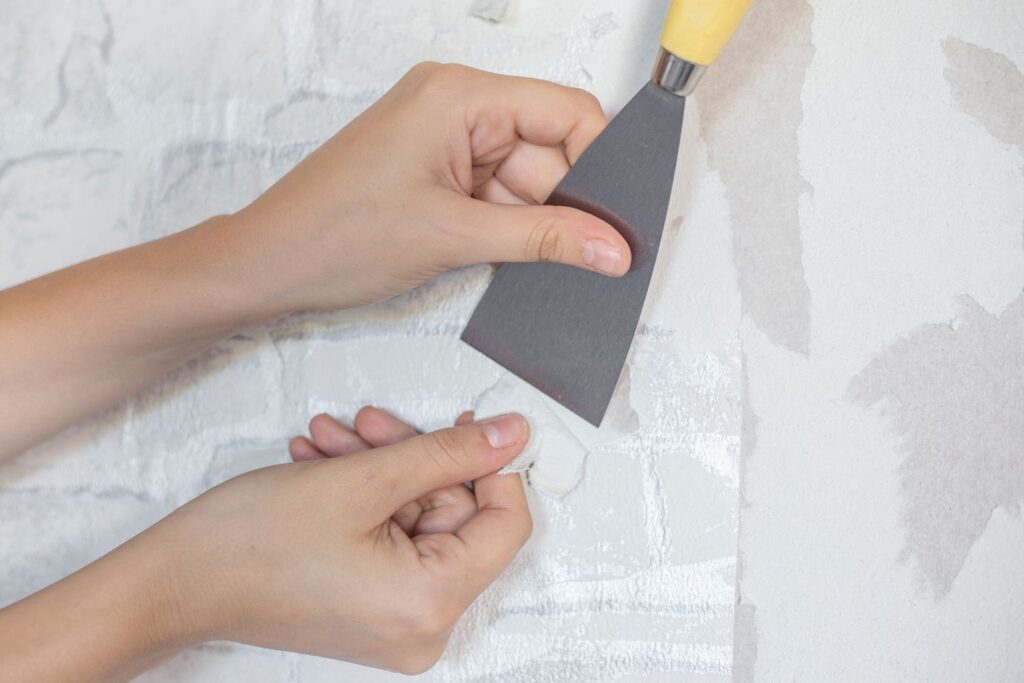 Hands using a metal putty knife to scrape old wallpaper off a textured white wall.
