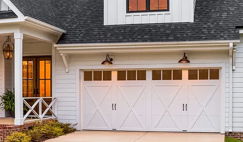 White farmhouse-style garage door with X-bracing and windows, next to a lit porch entrance.