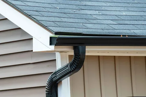 Black gutters and downspout on a house with a grey shingle roof and tan siding.