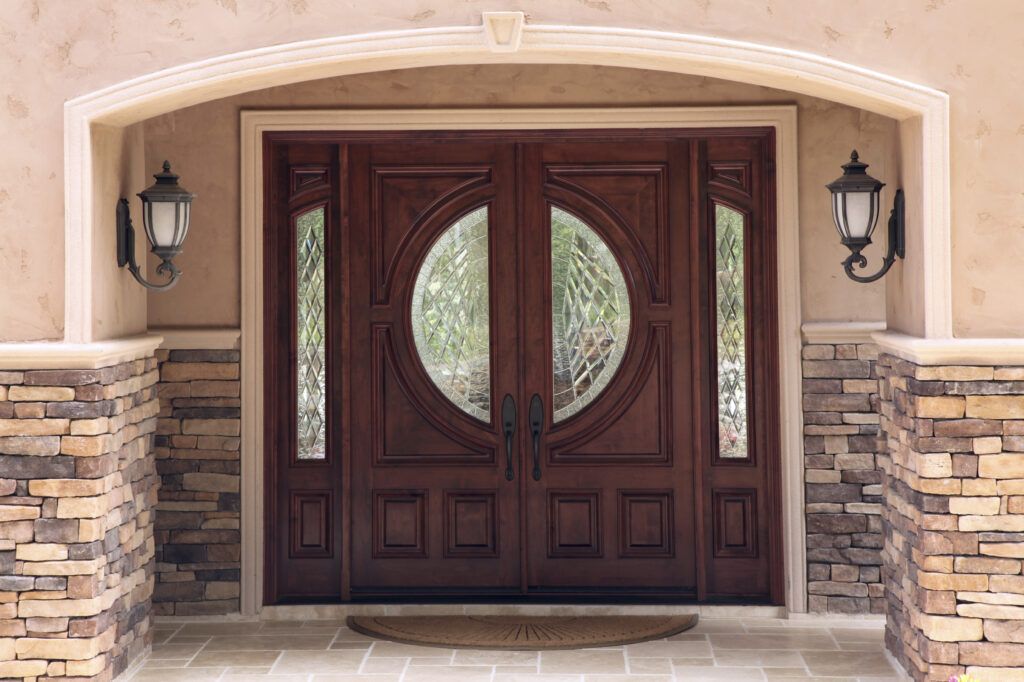Ornate dark wood double doors with circular glass inserts, flanked by stone pillars and wall lanterns.