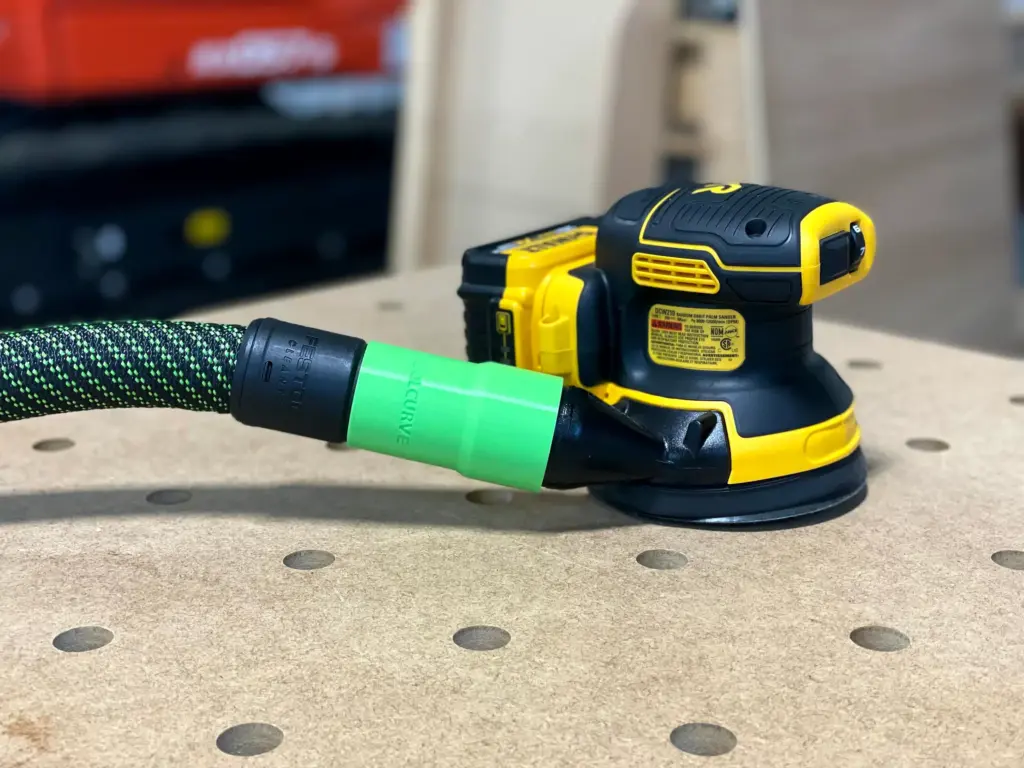 A yellow and black Dewalt random orbit sander connected to a Festool dust extraction hose on a workbench.