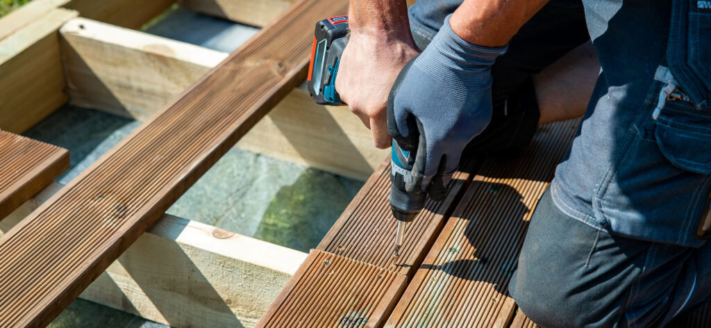 Worker using a cordless drill to install grooved wooden decking boards.