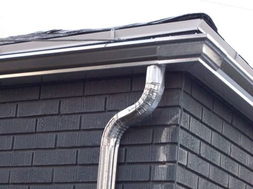 White gutters and a curved silver downspout on a grey brick wall.