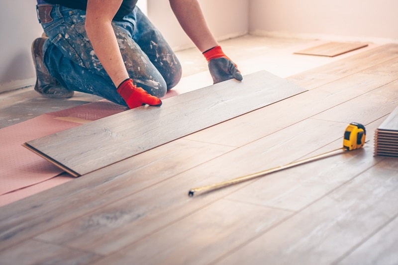 Worker in work gloves installing a wood laminate floor plank, with a tape measure nearby.