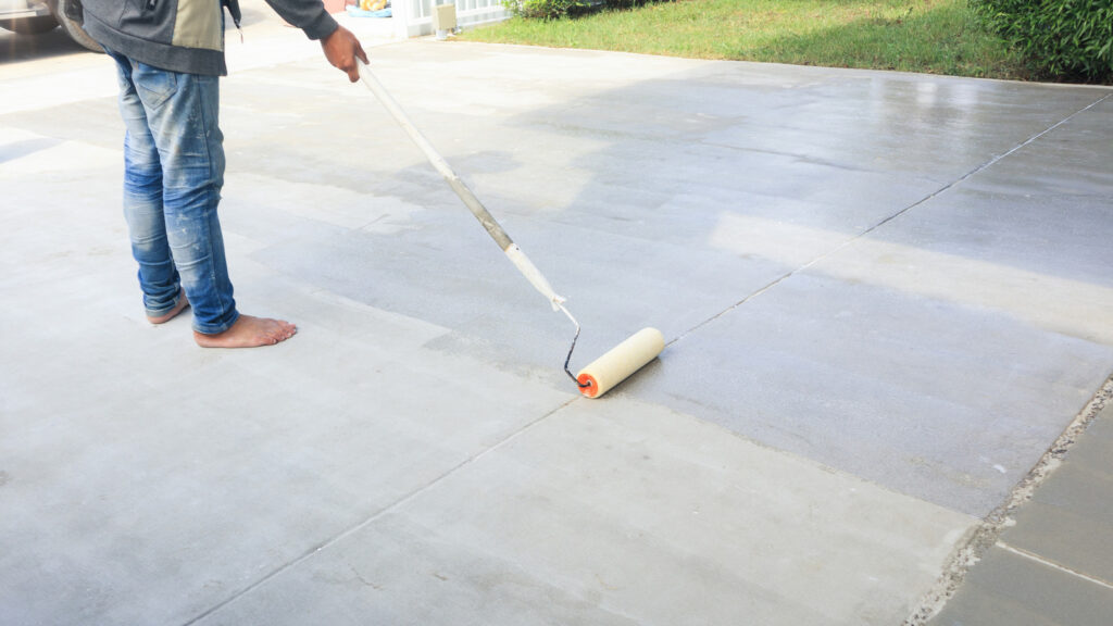 Person using a long-handled paint roller to apply sealant to a concrete patio.