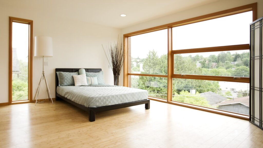 Minimalist bedroom with a low-profile bed, light blue bedding, and large windows overlooking a green neighborhood.