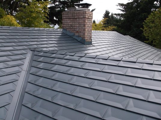 Grey textured metal roofing tiles on a house with a brick chimney and surrounding trees.