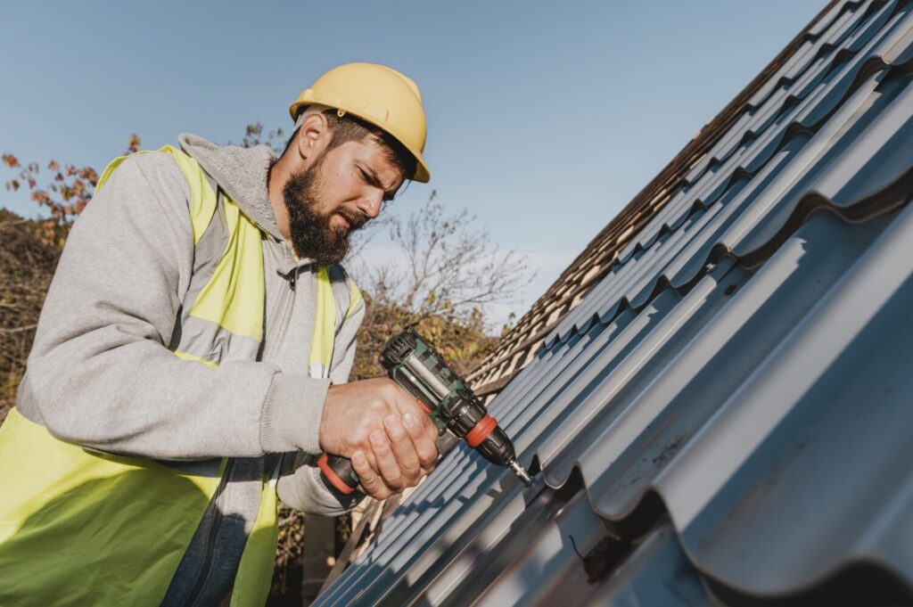 Construction worker wearing a hard hat and safety vest using a power drill on a metal roof.