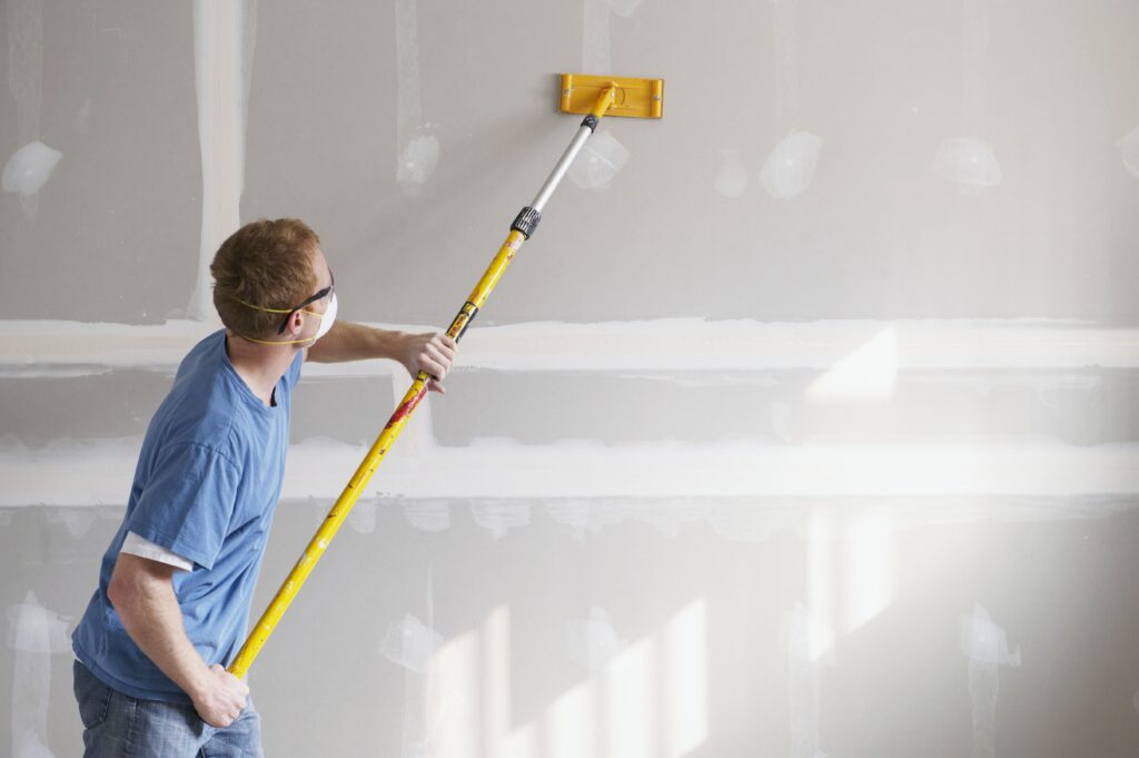 A person wearing a mask uses a long-handled sander to smooth a drywall wall.