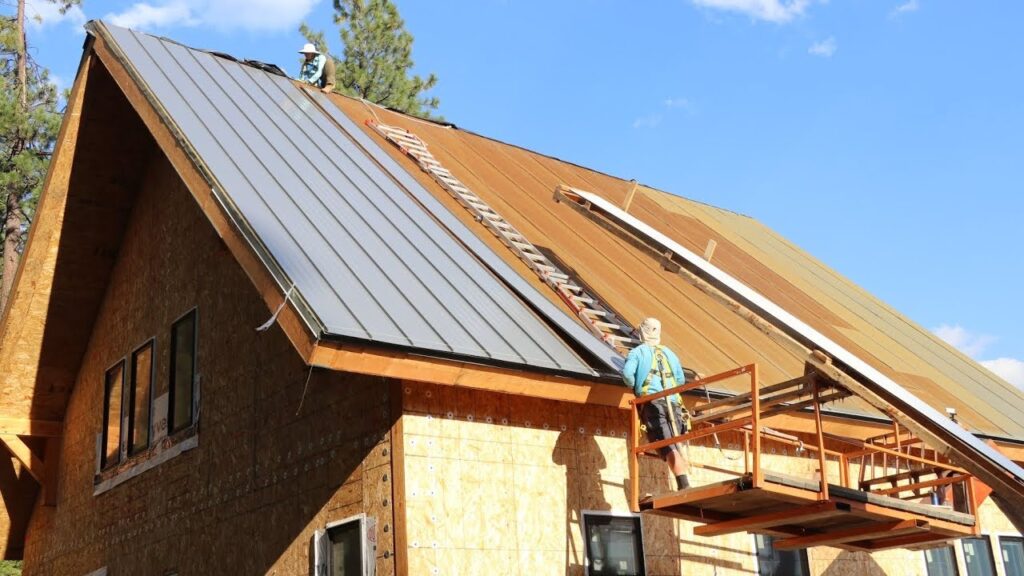 Workers installing gray metal roofing on a wooden house under construction.