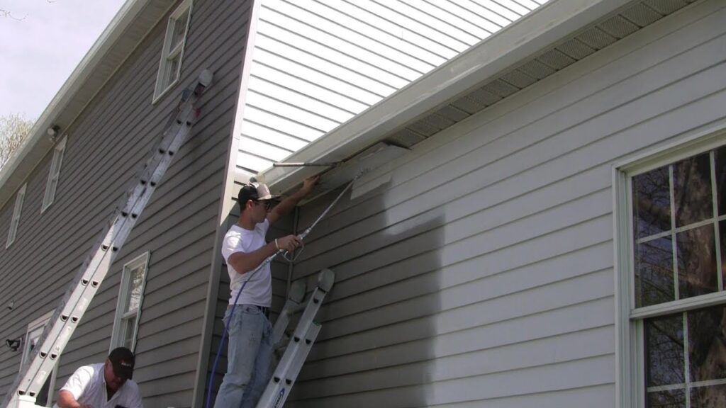 A man on a ladder uses a spray gun to paint the exterior gray siding of a two-story house.