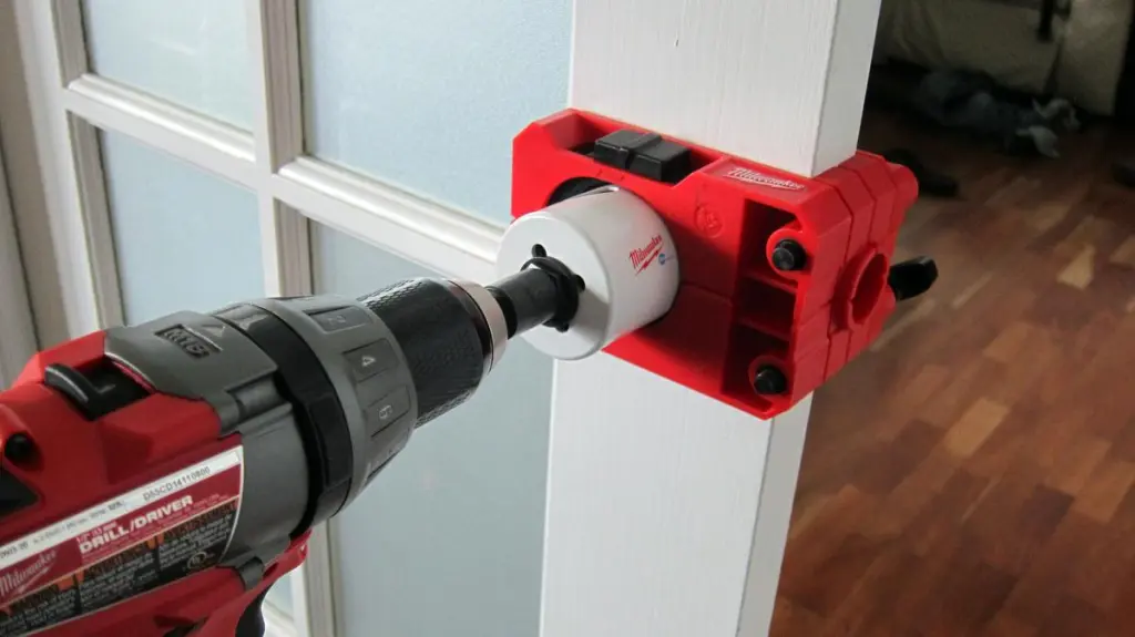 Milwaukee drill with a hole saw bit attached to a door lock installation jig on a white door frame.