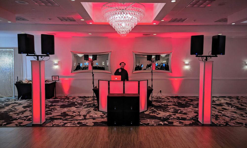 DJ behind a glowing white booth in an event hall with red ambient lighting and large speakers on stands.