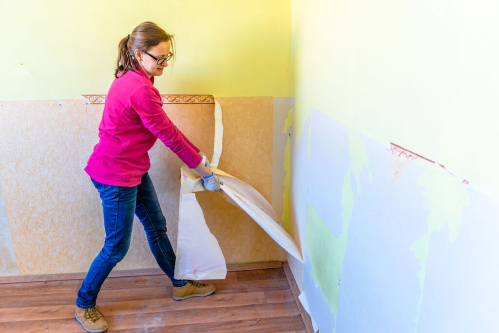 Woman in a pink shirt peeling old wallpaper off a wall during home renovation.