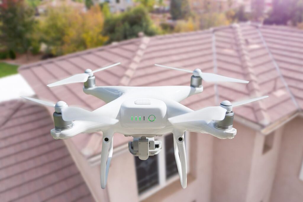White quadcopter drone hovering over a residential house with a tiled roof.