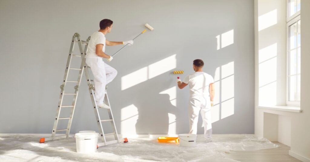 Two men in white clothes painting a gray wall, one standing on a ladder and the other on the floor.