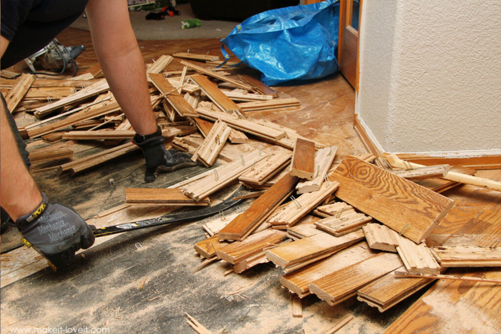 Person removing hardwood flooring with a pry bar, surrounded by ripped-up wooden planks and sawdust.