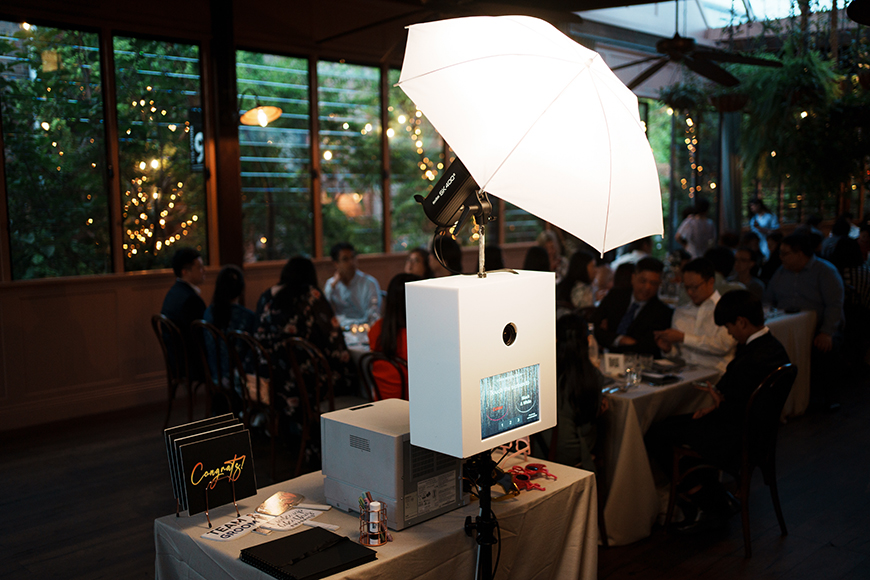 A white photo booth with a light umbrella stands on a table in a dimly lit room filled with guests at a banquet.