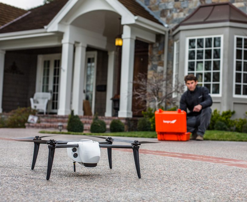 White drone on a driveway with a technician and orange equipment case in front of a large house.