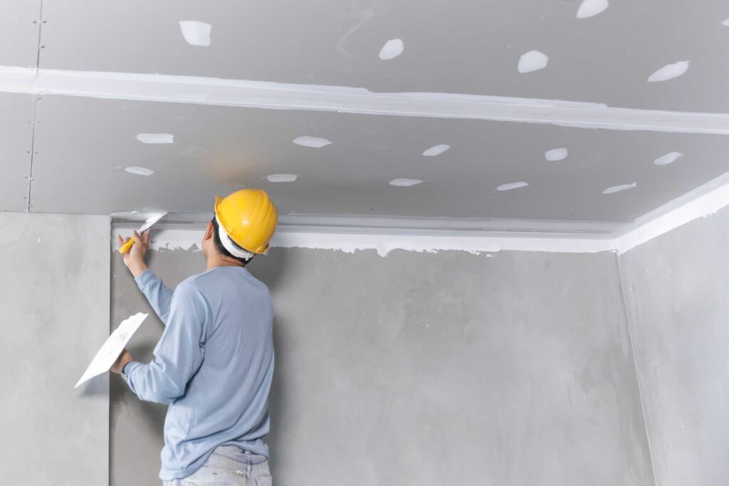 Worker in a yellow hard hat applying joint compound to a drywall ceiling with a trowel.