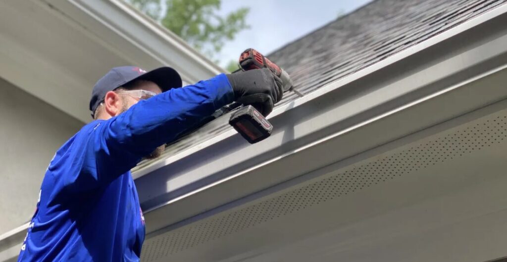 Worker in a blue shirt using a power drill to install white gutters on a house.